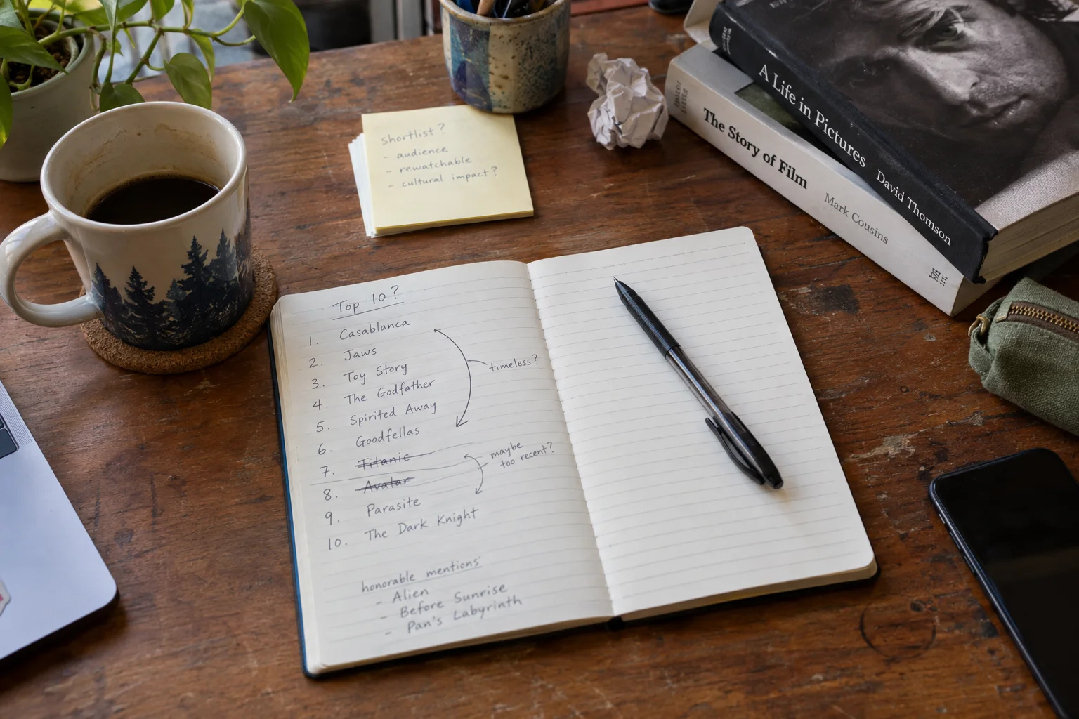 An open notebook of film rankings beside books and a coffee mug on a wooden table.